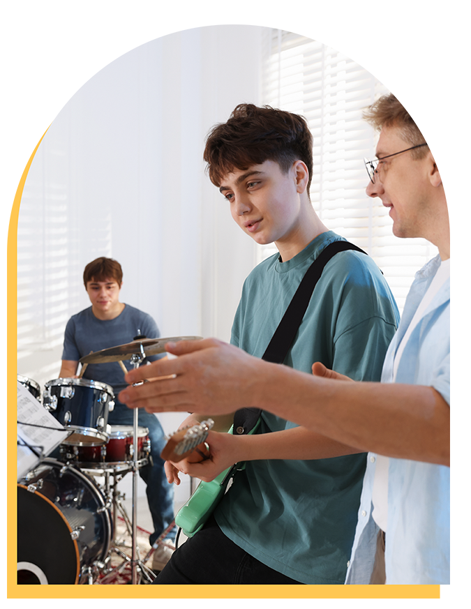 Three young people are playing music in a bright room; one plays drums in the background while two others stand in front, one holding an electric guitar and the other gesturing with his hands as they interact.
